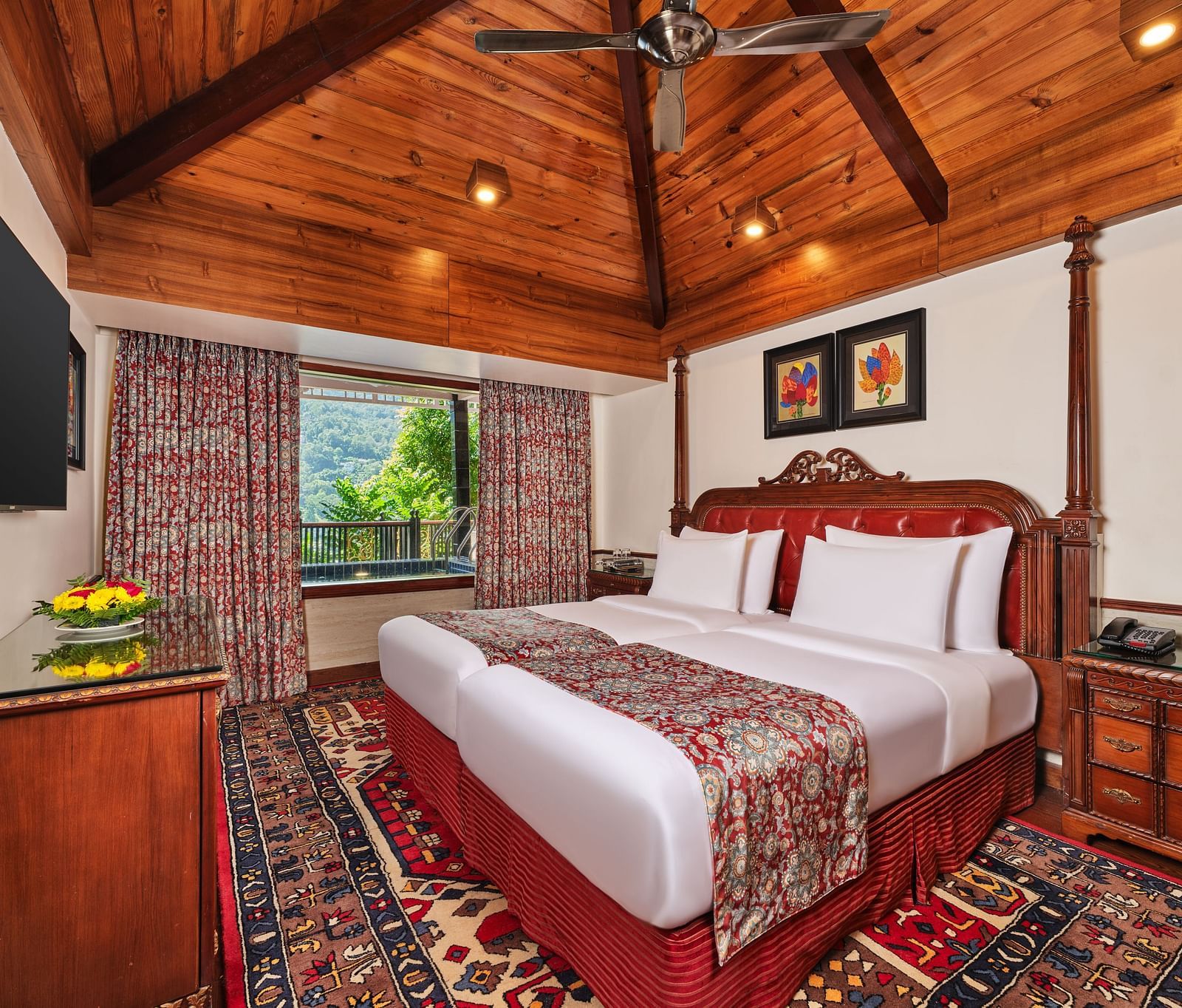 Imperial Villa bedroom with a large bed and decorative rug, a table, wooden cabinet beneath a TV , wooden ceiling and a window with decorative curtains overlooking greenery