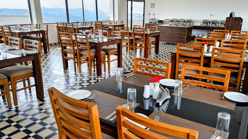 Seating arrangement inside the in-house restaurant serving the best food in Vagamon, with floor to ceiling windows in the distance having valley-views at Abad Silvermist, Vagamon