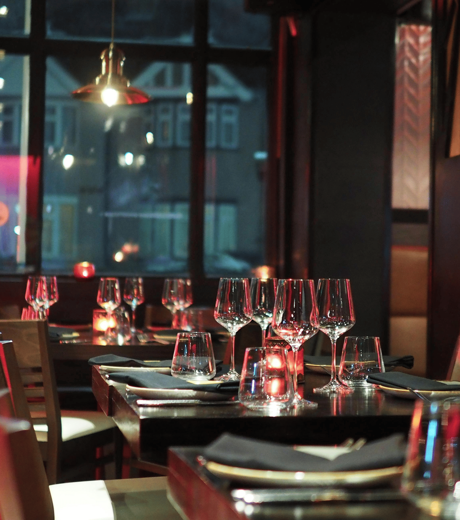 A moody interior shot of a restaurant dining table set with wine glasses and candles, seen through a dark window.