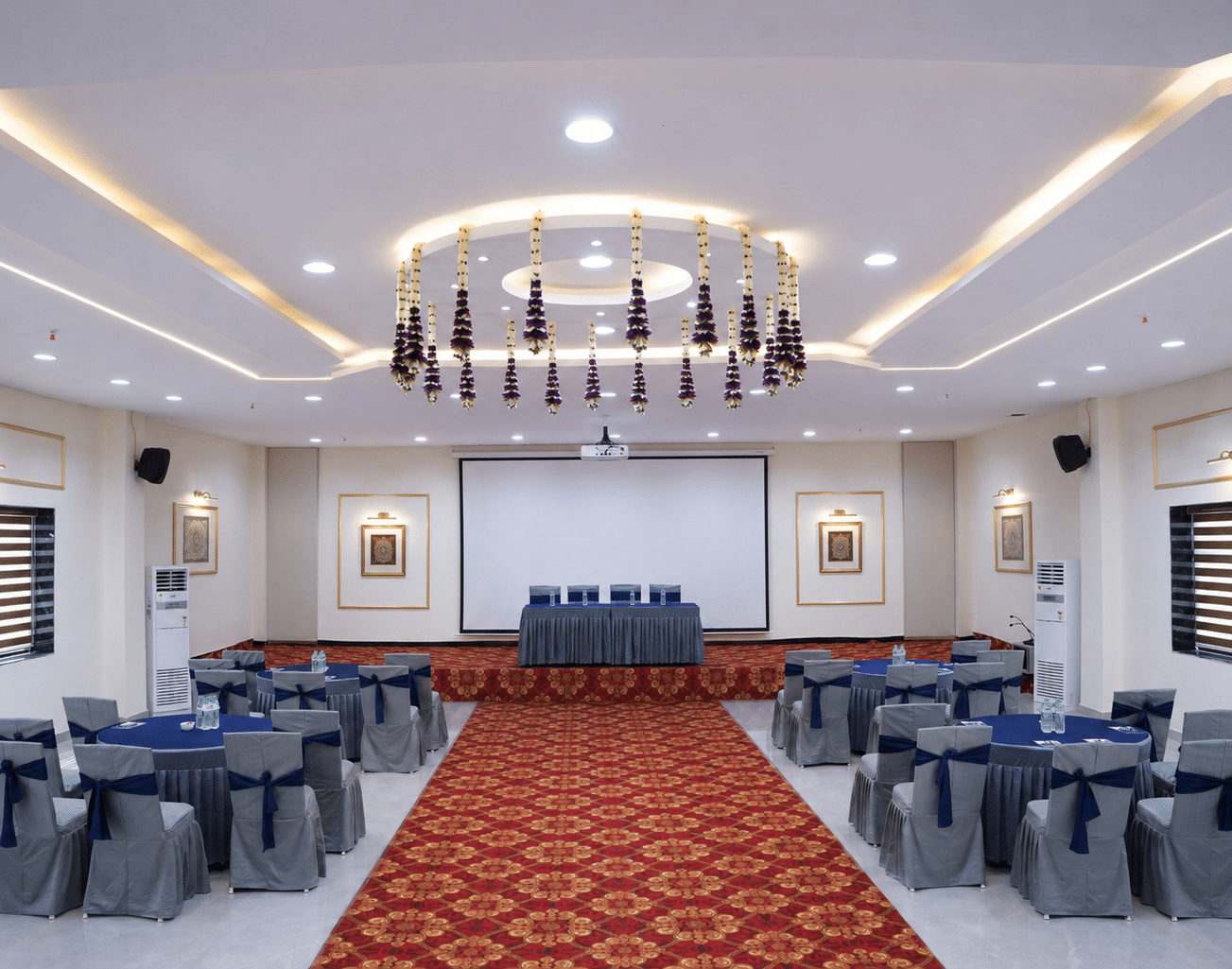 The large banquet hall setup of Galadari Boardroom at Hotel Galadari with round dining tables, chairs, a main stage, and overhead floral decorations.