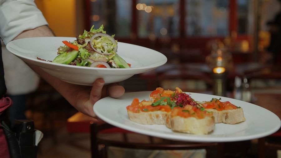A waiter in a white shirt and black waistcoat carries two plates, one with a salad of lettuce, onions, cucumbers, and tomatoes, and another with bruschetta.