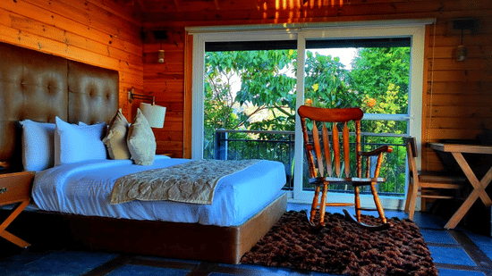 Wooden cabin room at Polo Resort Cherrapunji with tufted leather headboard king bed, polished rocking chair, side desk and sliding doors to greenery.