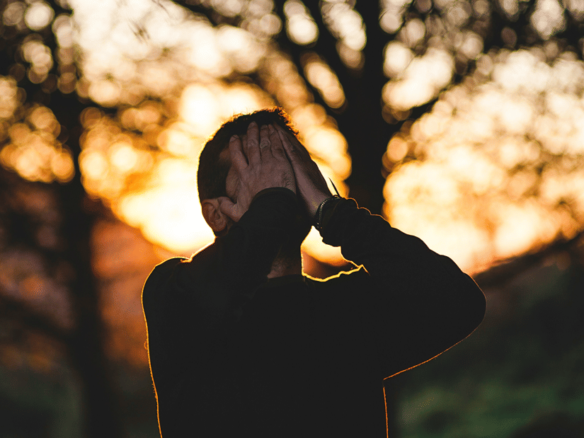 A man silhouetted against sunlit trees, holding his head in both hands