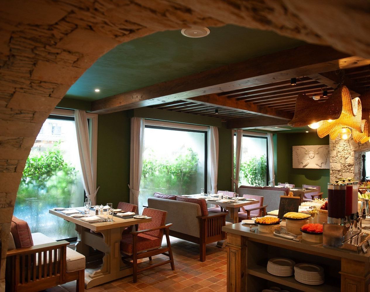 A wide-angle view of the dining area at Mila Restaurant at The Evren, Vagator, captured from a stone archway. The space features wooden tables, comfortable chairs and banquettes, and a buffet station with a variety of food.