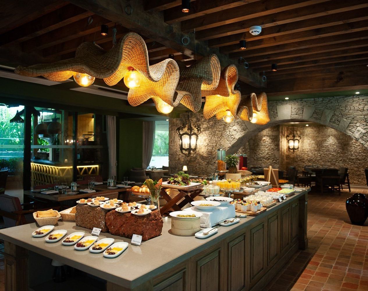 A lavish breakfast buffet spread at Mila Restaurant at The Evren, Vagator, featuring a variety of dishes on a large central island. The restaurant has a rustic yet modern ambiance with woven light fixtures, a stone archway, and a view of the outdoors.