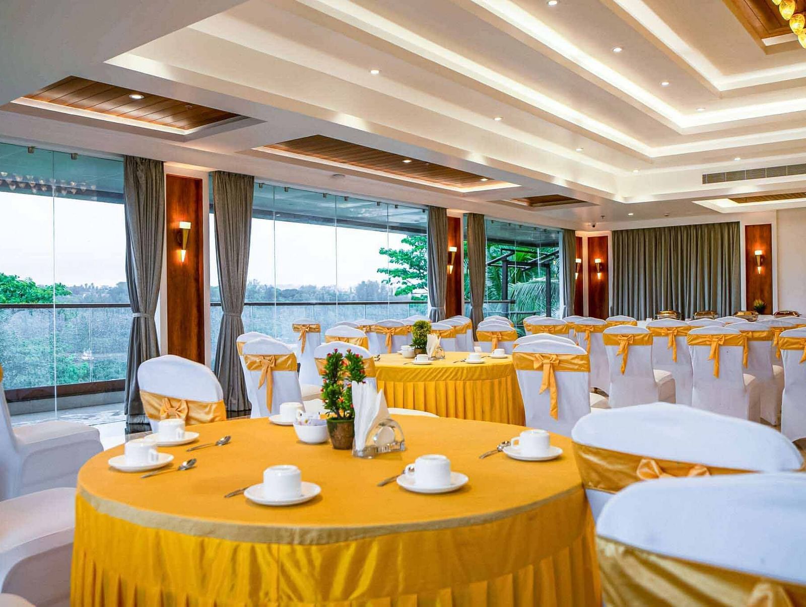 A bright and airy event hall at Mansha Palace is set up with round tables draped in yellow and white chairs with yellow sashes, offering a pleasant view through large windows.