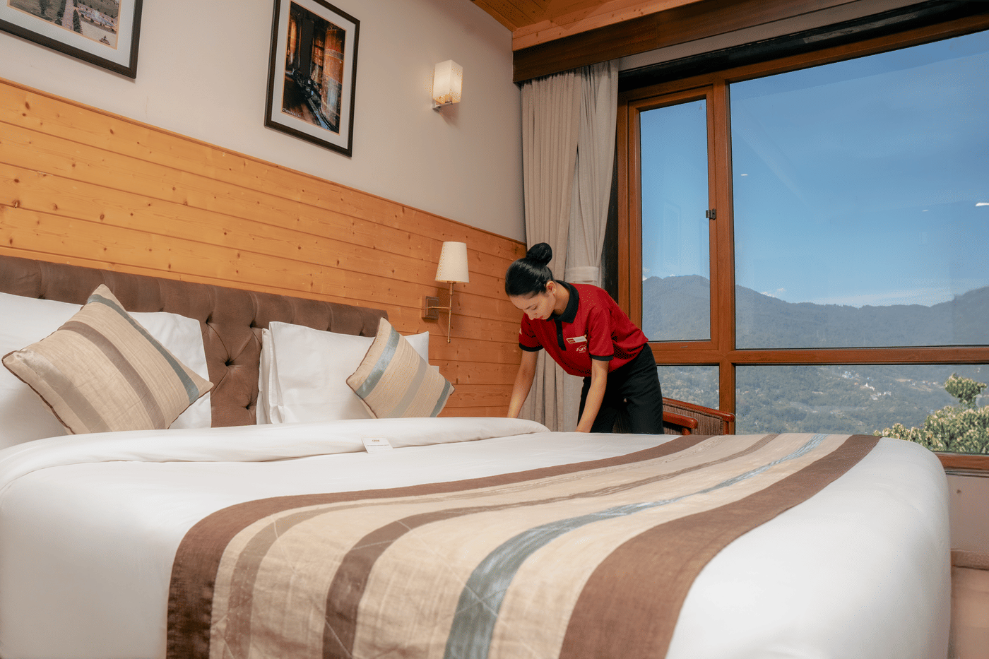 Hotel staff in uniform neatly making a bed with crisp linens in a cosy room featuring wooden walls, large pillows, and a scenic mountain view through a window.