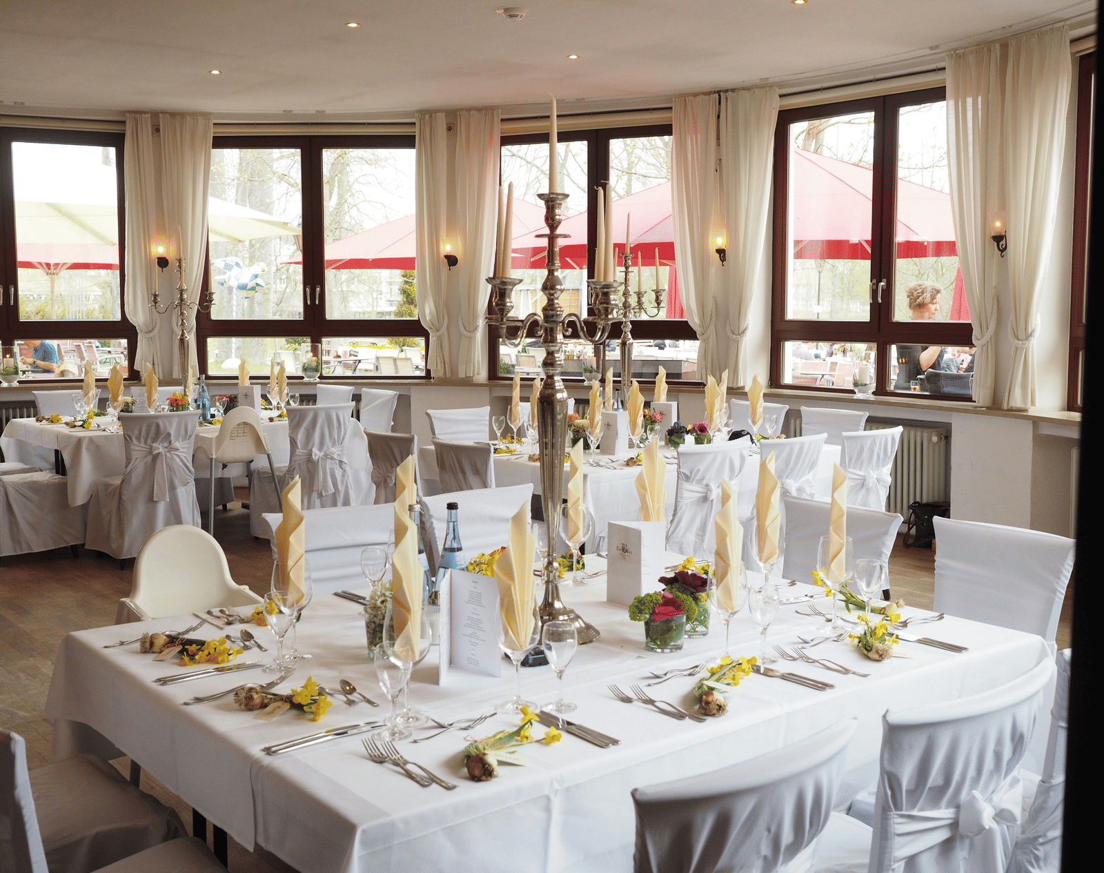 A dining room set up with multiple tables covered in linens and chair covers, featuring a central square table with a tall candelabra, cloth napkins, and windows in the background.