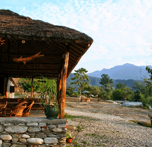 image shows a thatched-roof tent stay at the jamoon corbett, with a view of the mountains in the background.