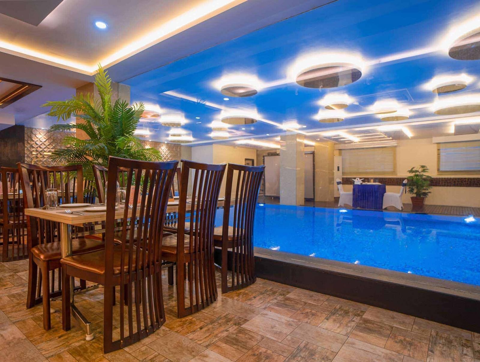 A dining area at Mansha Hotel Regency with a distinctive blue-lit ceiling, dark wooden tables and chairs, and a pool in the foreground.