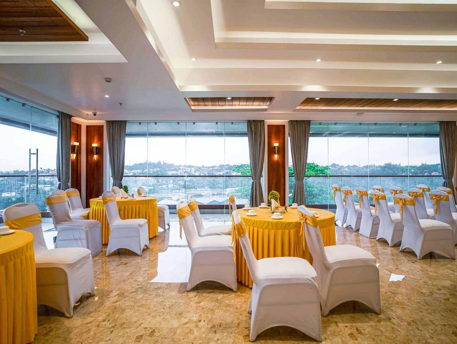 A bright event space at Mansha Palace offers city views through large windows and is furnished with set tables and chairs.