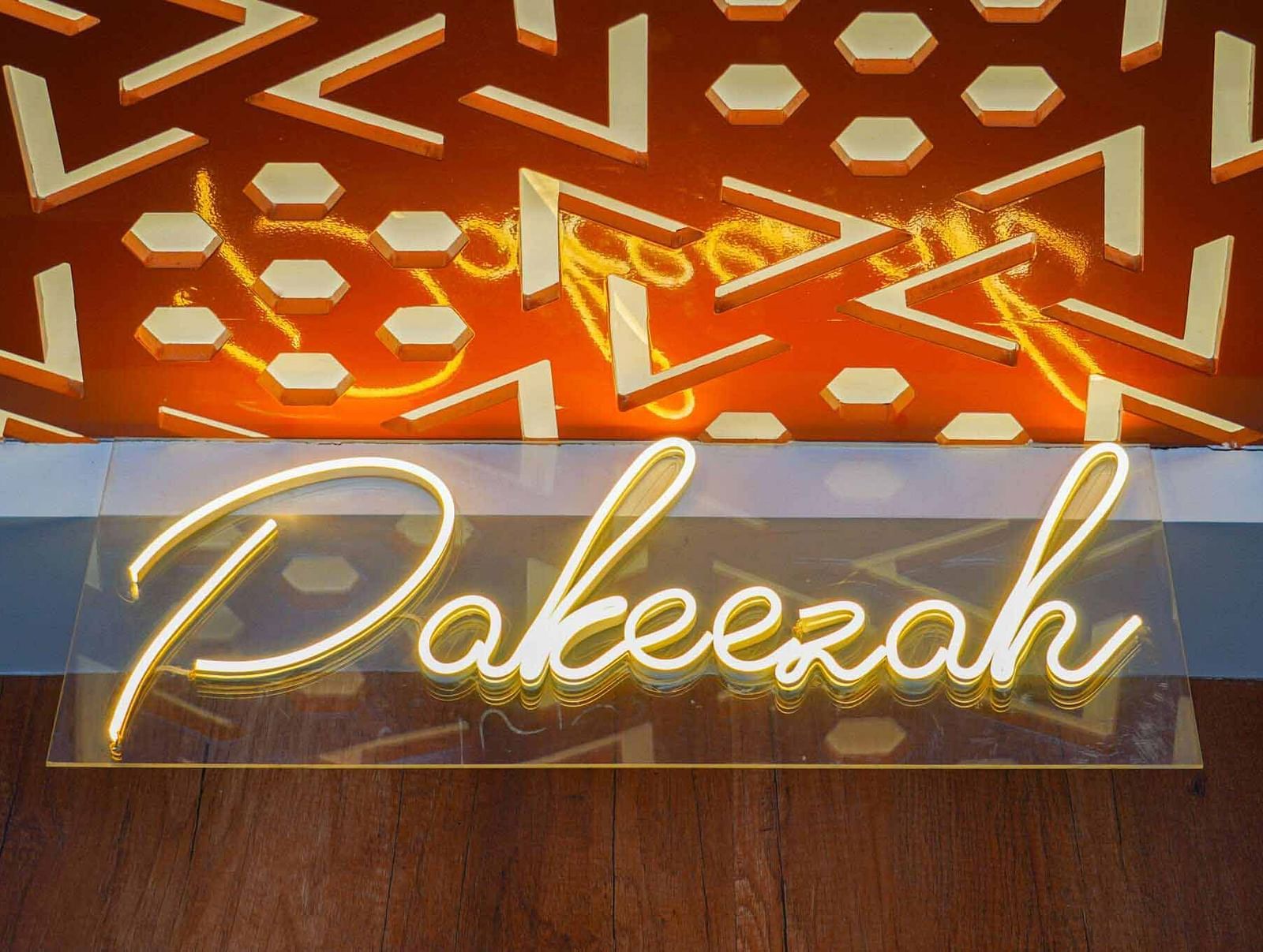 A glowing neon sign displaying Pakeezah in a cursive script is mounted on a patterned wall.
