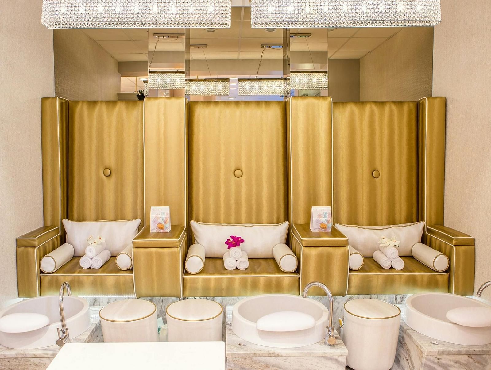 Spa pedicure setup with three plush gold chairs, each with towels and pillows, facing foot basins and a white countertop.