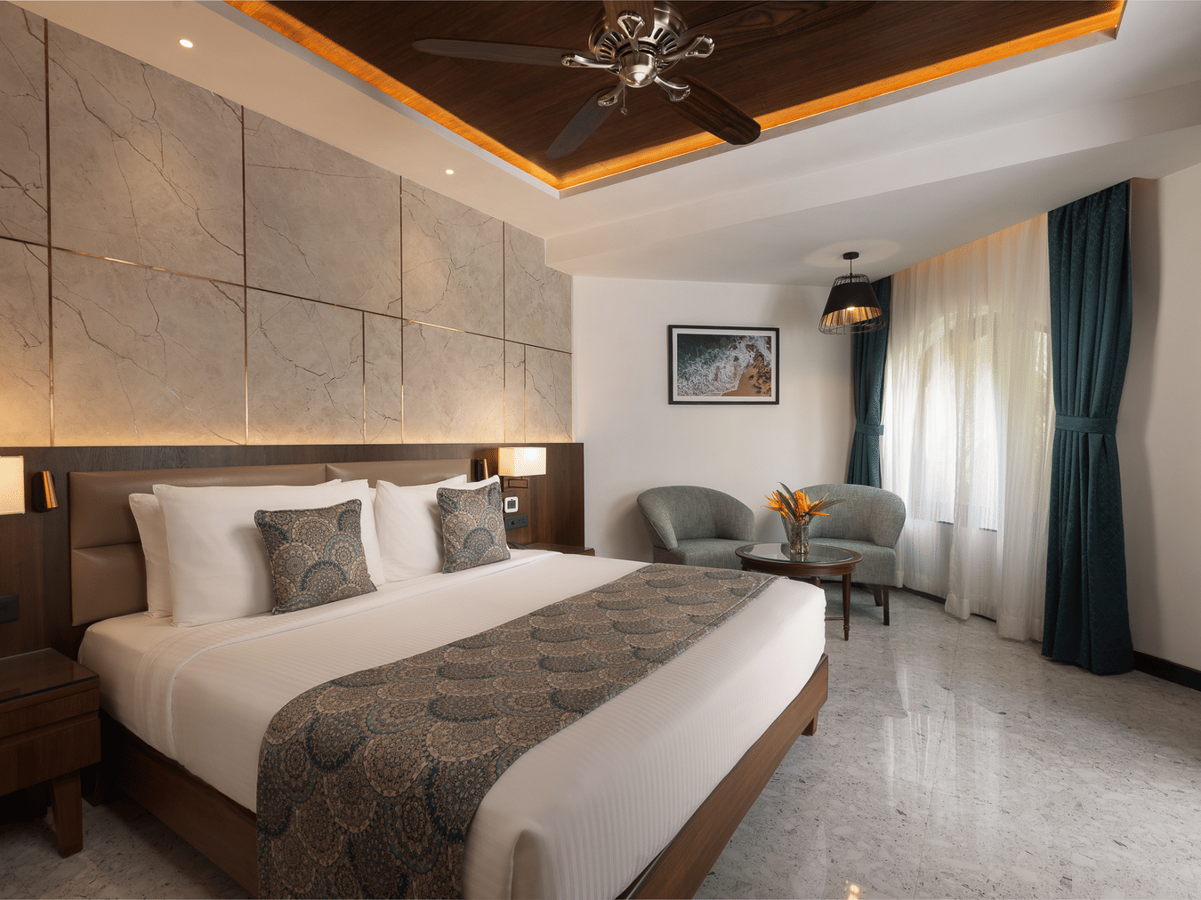 A view of the Junior Suite with a king bed, side tables, side lamps, ceiling fan and chairs in the corner at Kenilworth Resort & Spa, Goa.