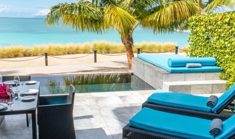 Beachfront patio with turquoise sun loungers, palm trees, and a dining table set against the backdrop of golden sand and calm Caribbean waters.