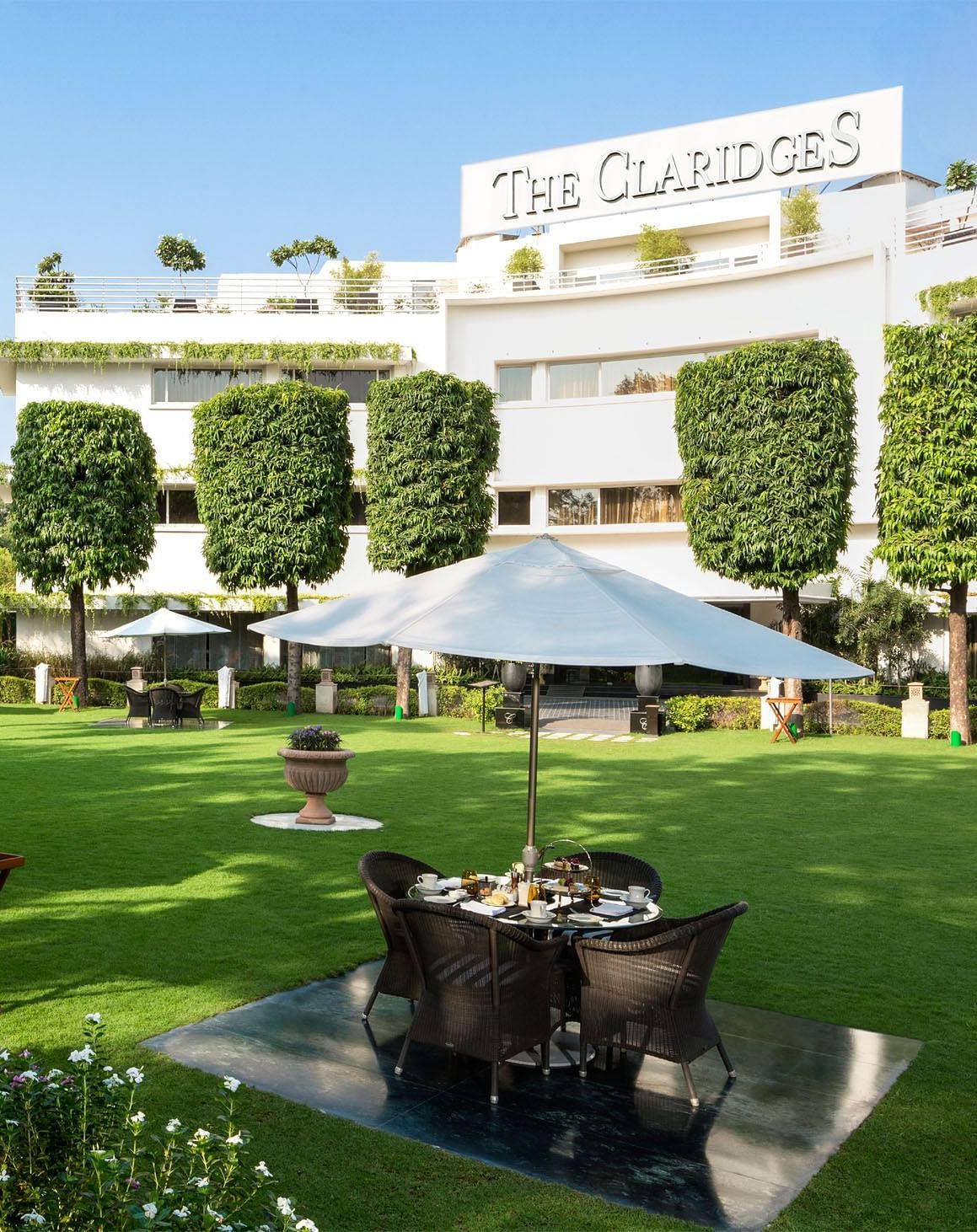 Facade of The Claridges New Delhi, featuring a green garden in the front and surrounded by towering trees, offering a serene and elegant ambience