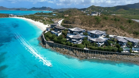 An aerial view shows a luxurious resort nestled along a pristine turquoise coastline with lush green hills.