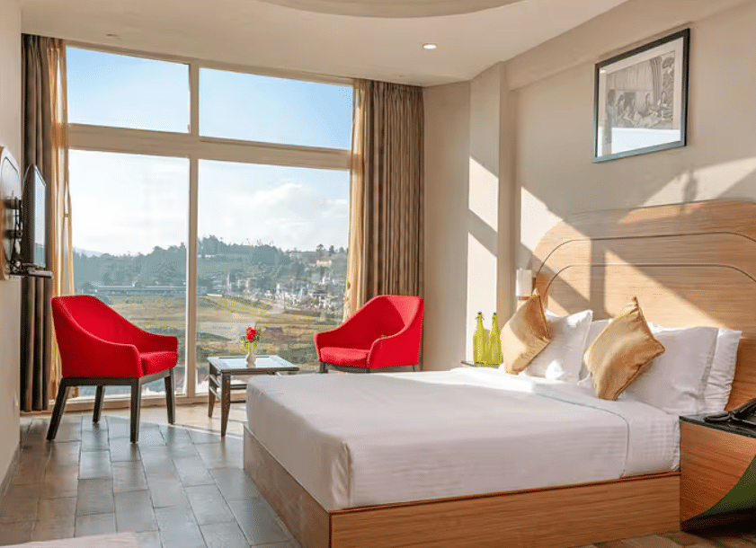 A hotel room with a large bed, two red armchairs, a side table, and a panoramic window overlooking a scenic landscape.