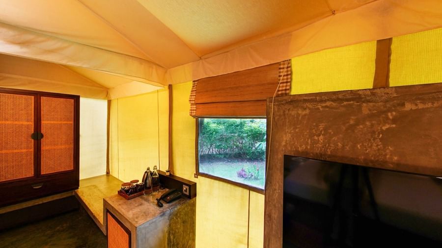 Wooden cabinet, stone counter with bottled water and tray, telephone, and window with garden view inside a yellow fabric-walled tent at Fireflies Resorts, Kabini.