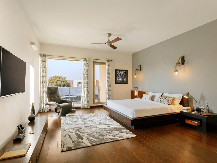 A spacious bedroom with dark wood floors, a low-profile bed, a large window leading to a balcony, a ceiling fan, and a wall-mounted TV - Karma Lakelands