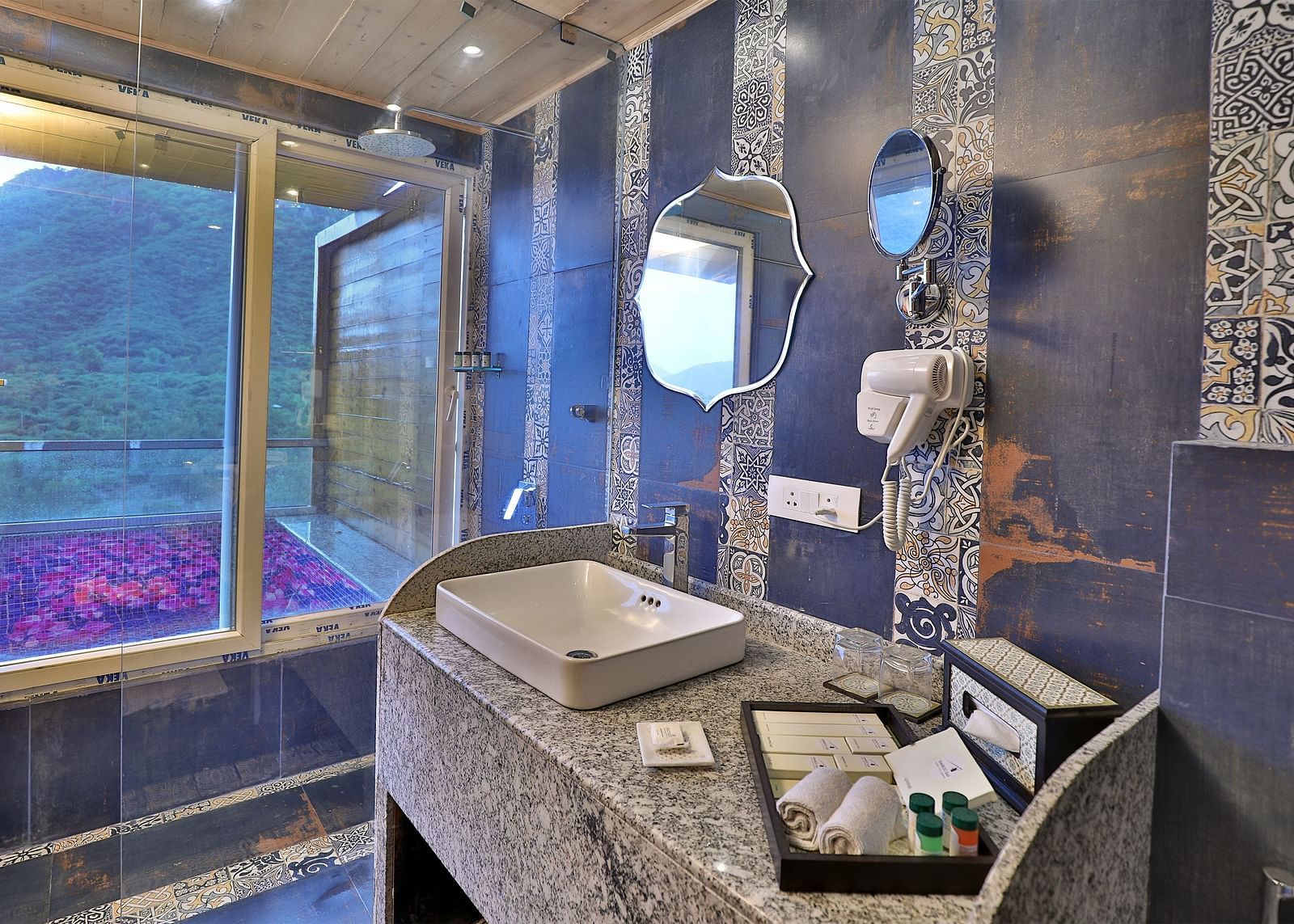 A bathroom with a unique patterned tile design, a sink on a speckled counter, and a window overlooking a lush landscape.- Bamboo Saa Resort & Spa, Udaipur