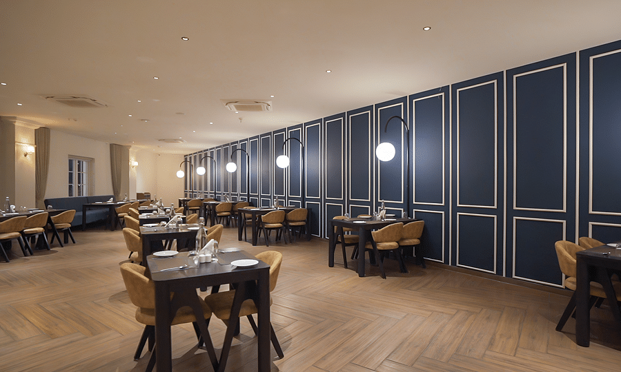 A well-lit restaurant dining room at Pride Resort in Rajkot featuring wooden tables, chairs, and a striking dark blue paneled accent wall, creating a modern and inviting atmosphere
