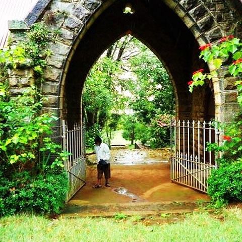 An arched stone entrance covered in greenery, leading to a path with a person walking through it.