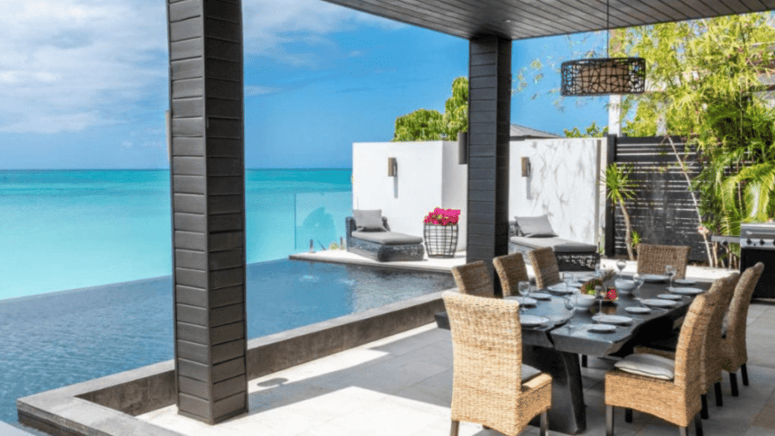 A luxurious outdoor living space with a dining table and chairs sits beside an infinity pool, all facing a serene turquoise sea.