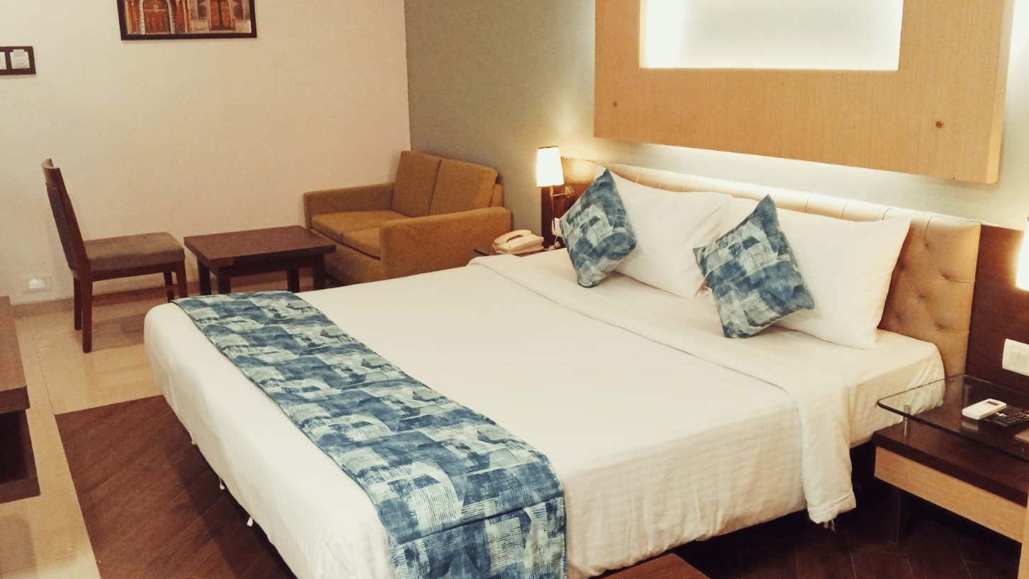 A bedroom layout showcasing a king size bed, a fabric sofa, a single chair, a wooden coffee table, and a framed wall painting at Hotel Mumbai House, Thane.