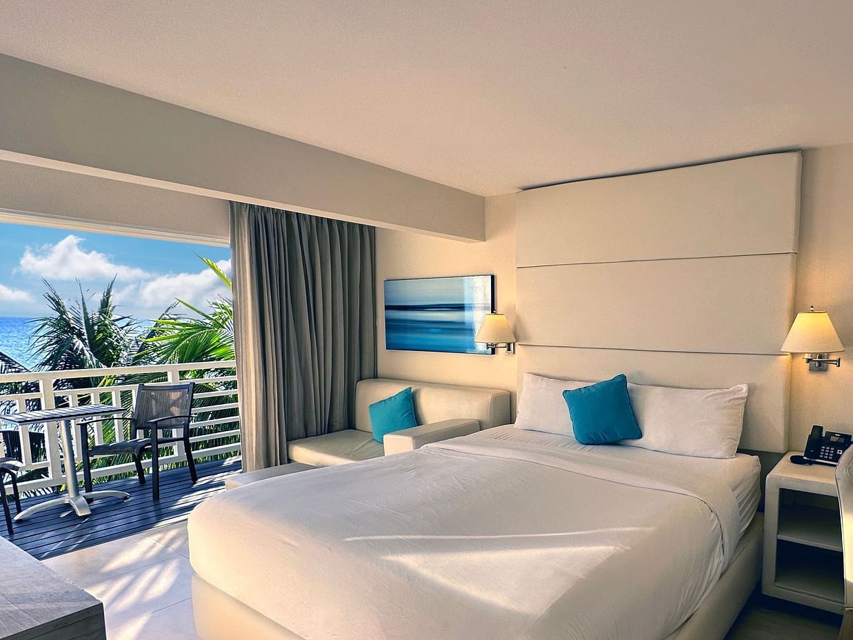 A bright hotel room with a large bed, sofa, desk, and balcony overlooking palm trees, featuring modern decor and teal accents.