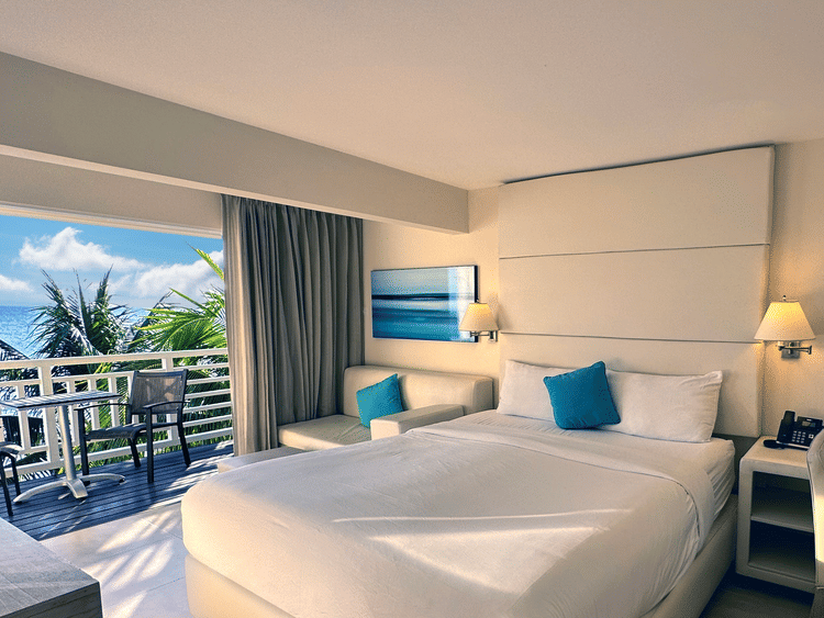 A bright hotel room with a large bed, sofa, desk, and balcony overlooking palm trees, featuring modern decor and teal accents.