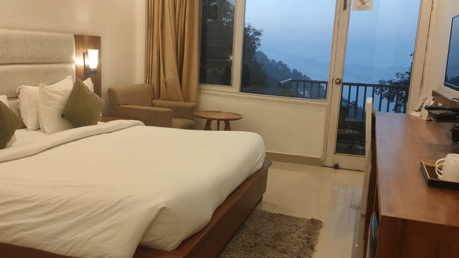 A room at Pride Ashiyana Resort Mussoorie with a bed, TV, desk, chair, and a balcony showing a mountain view.