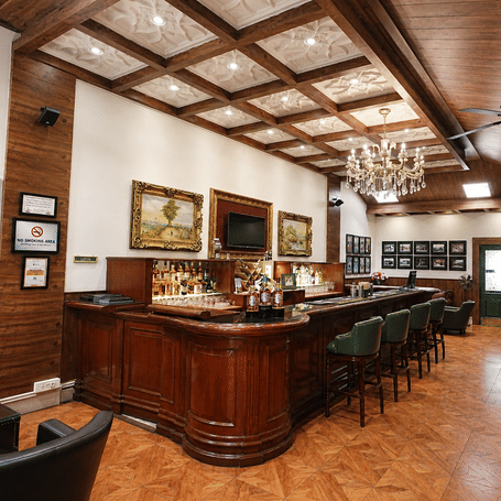 John's Tavern bar with a polished wooden counter, green leather stools, a crystal chandelier and framed vintage prints at MAYFAIR Himalayan Spa Resort, Kalimpong.