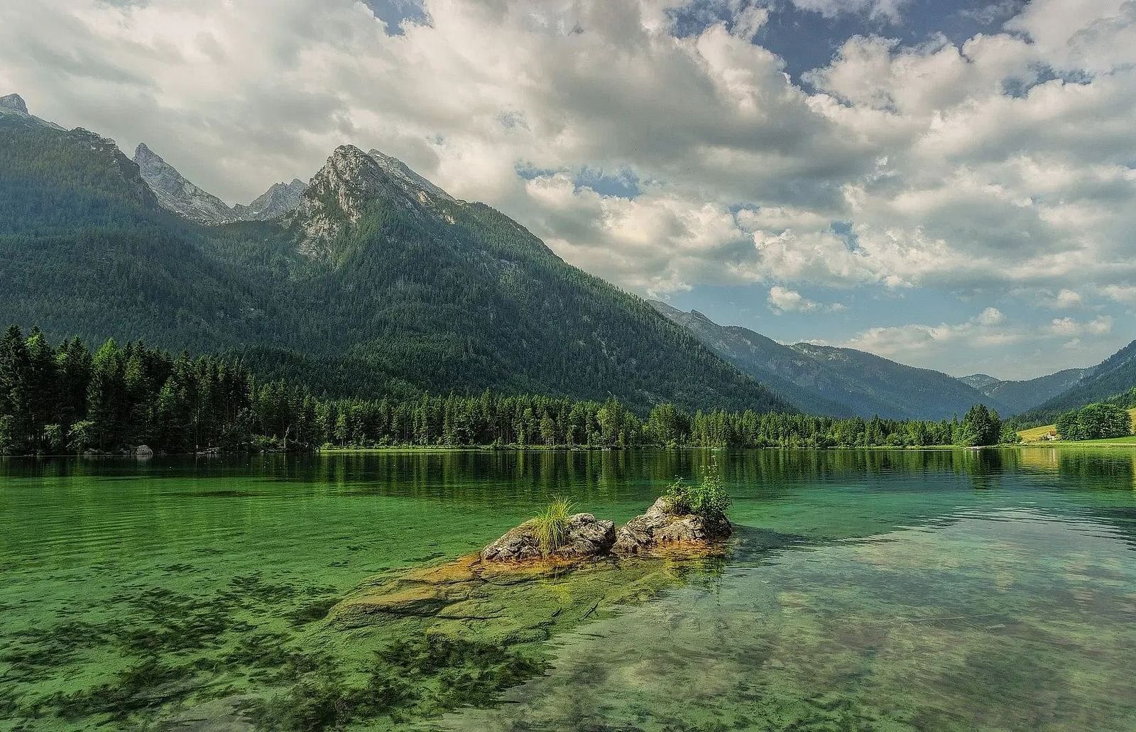 Lake reflects mountains, trees line the shore, cloudy sky above.