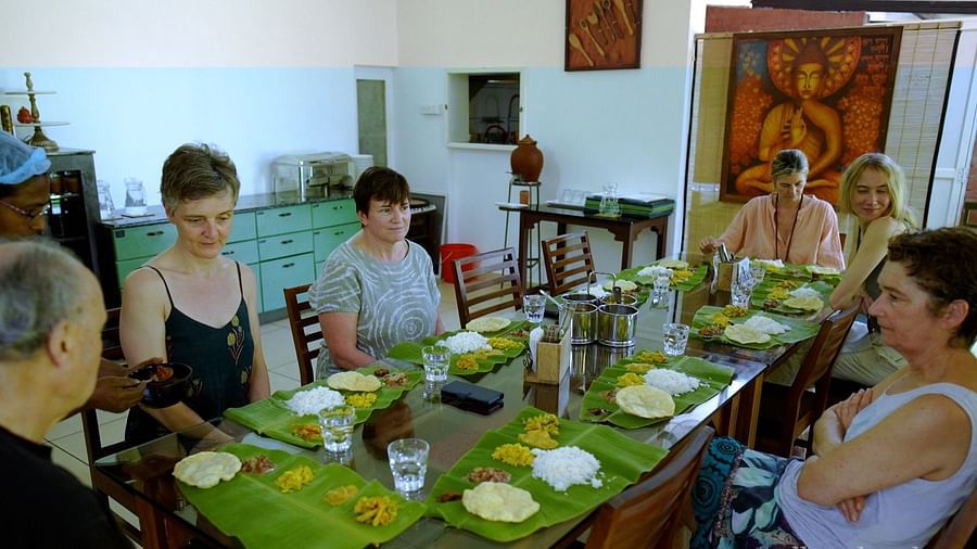 A group of people are seated at a long table indoors, sharing a meal served on banana leaves.