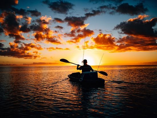 a person kayaking on a water body with the sun setting in the background making the sky turn into hues