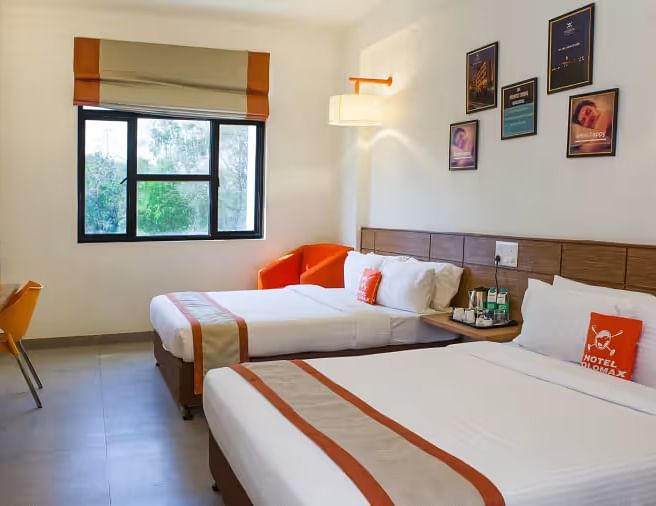 View of a hotel room featuring two comfortable beds, wall art, and a large window with natural light - Max Hotel Prayagraj.