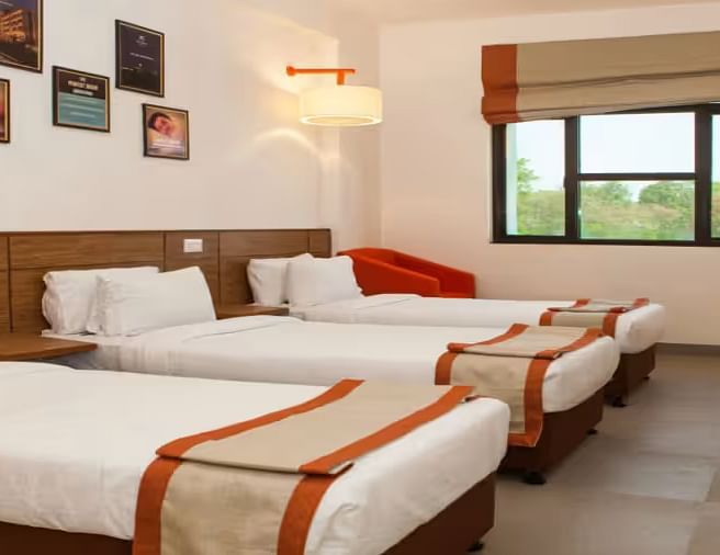 A bright, well-furnished hotel room with three neatly made beds, wall art, and a window- Max Hotel Prayagraj.
