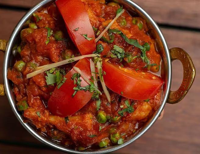 A bowl of Indian curry with visible peas and paneer, topped with tomato slices and cilantro, in a brass serving dish.
