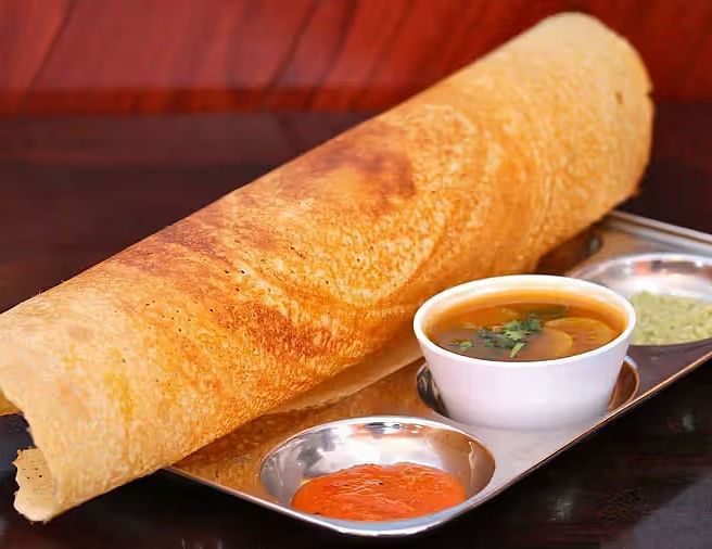 A close-up of a crispy dosa rolled and served with a bowl of sambar and two colorful chutneys on a metal plate.