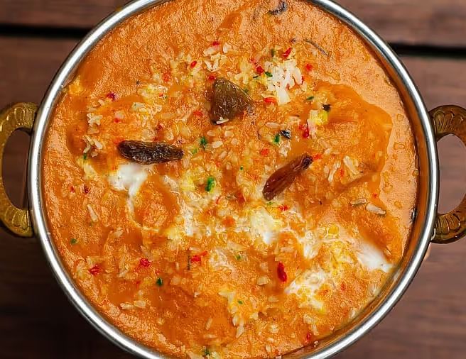 A close-up of a rich, creamy orange Indian curry with visible textures and colourful garnishes in a traditional brass serving bowl.