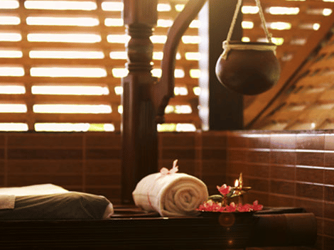 A pot hanging from a wooden bar over the spa bed next to which is a rolled-in towel and flowers at Ayur Ashram, Uday Suites
