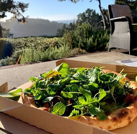 Arugula-topped pizza in an open box on a patio with a scenic view in the background.