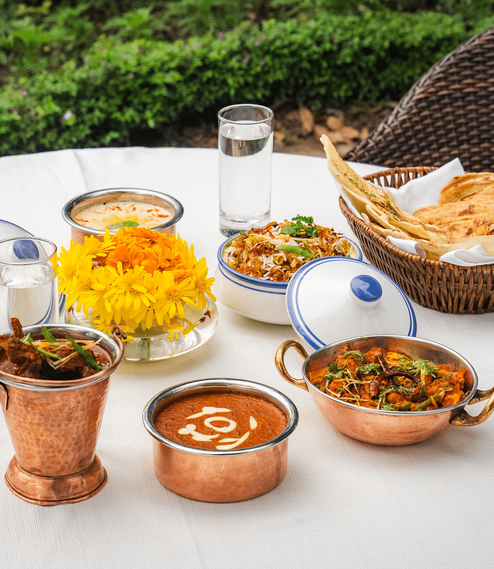 An image of a table with various Indian dishes in copper pots, naan bread, water glasses, and flowers.