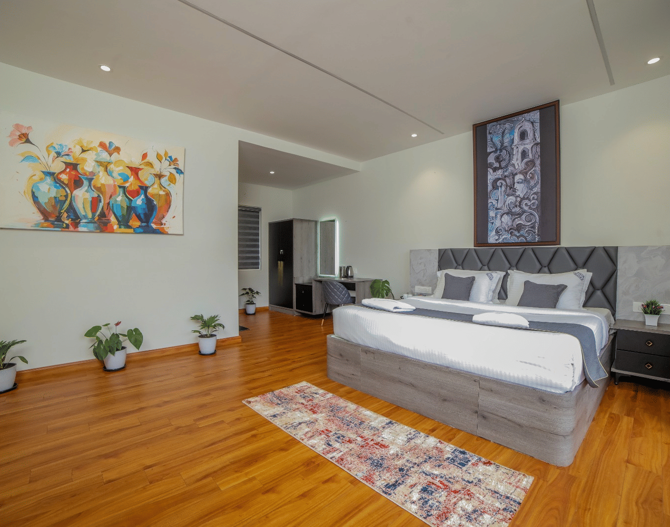 A wide view of a well-lit bedroom in MYST Resorts, Doddabetta, Ooty, featuring wooden flooring, a plush queen size bed, paintings hung on the wall, and one corner of the room where furniture is placed.