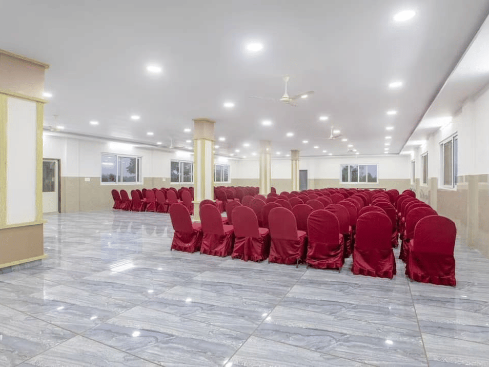 Back view of a spacious banquet hall at Siri Ambari Resorts and Spa, featuring red-covered chairs and bright lighting.