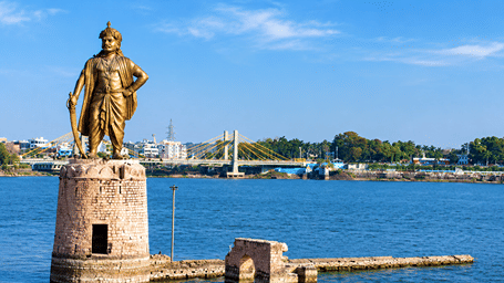 Golden statue of Raja Bhoj standing tall on a stone pedestal over the waters of Upper Lake in Bhopal with a cable-stayed bridge behind.