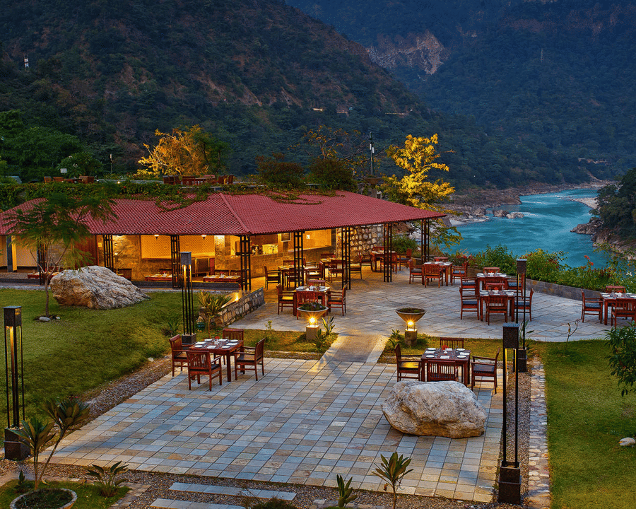 Outdoor dining area by a river, with tables set on a stone patio. A red-roofed pavilion is surrounded by lush greenery and mountains, conveying tranquility - Aloha On the Ganges