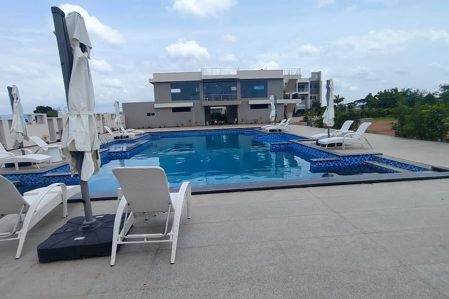 Rooftop swimming pool with tiled edges, surrounded by lounge chairs and closed umbrellas; a modern building is visible in the background, with trees and open space to the right. A person is partially visible in the foreground on the left - Kyriad Prestige,