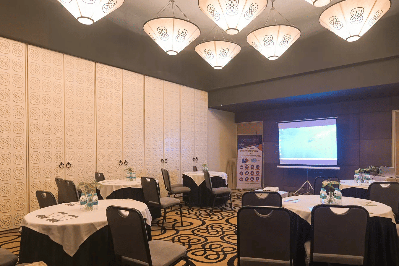 Round banquet tables set up for a seminar facing a projector screen in Dholavira banquet hall at 3102bce - A Vedic Resort by Lindsay.